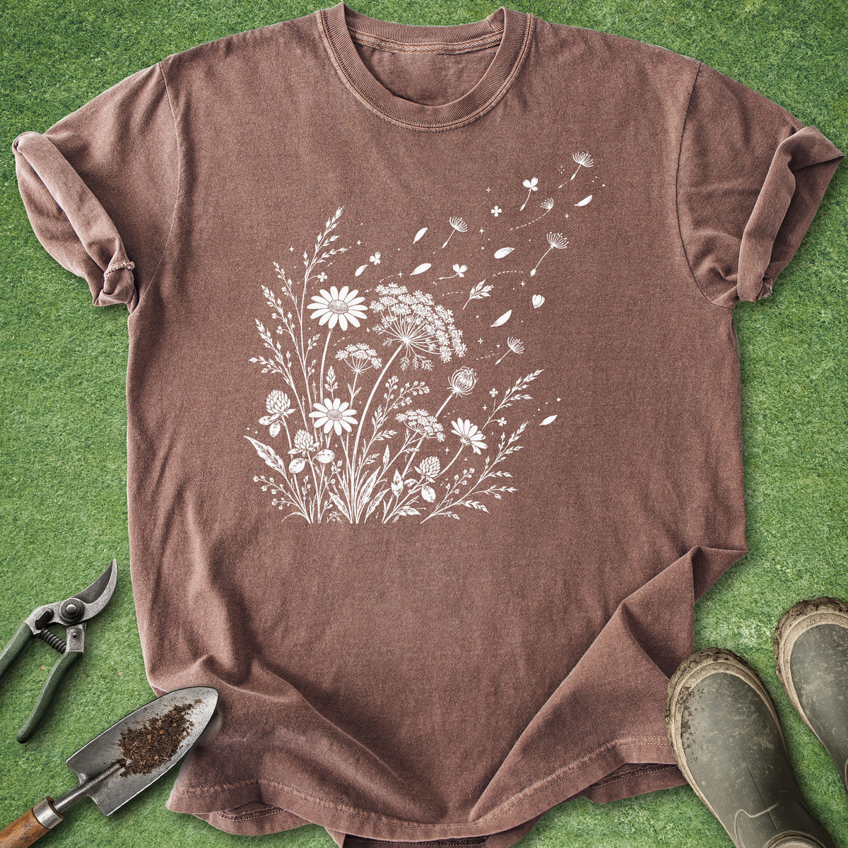 A brown short-sleeve crewneck t-shirt with a white wildflower graphic on the front, laid flat on green grass with gardening props including pruning shears, a trowel, and boots.