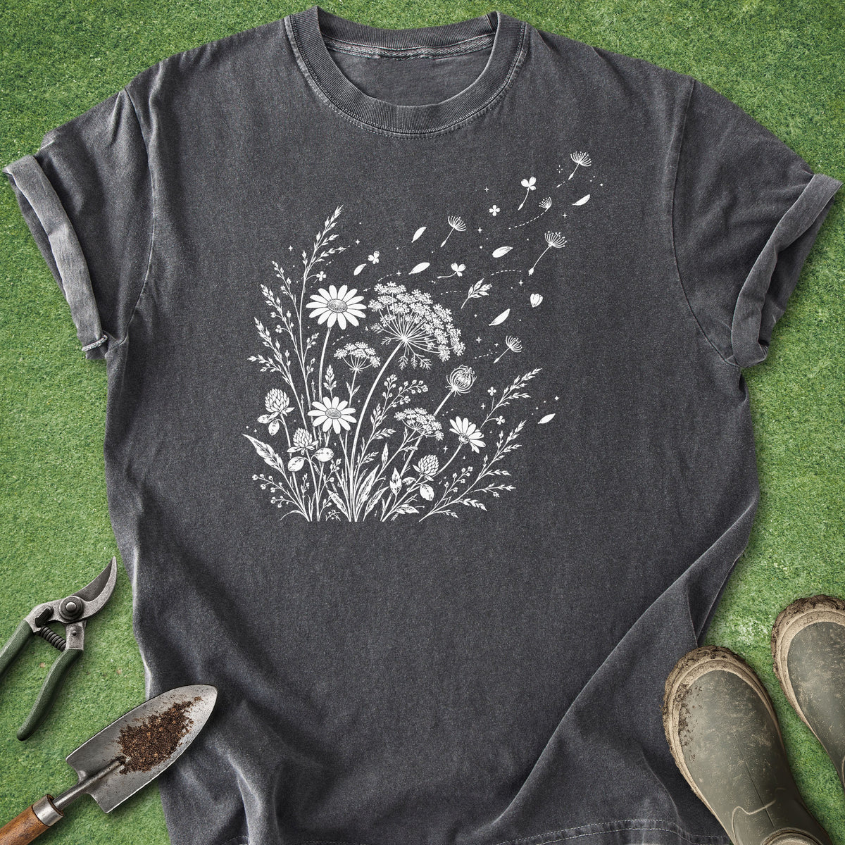 A dark gray t-shirt with a white wildflower graphic print, styled flat-lay on green grass with gardening tools and boots.