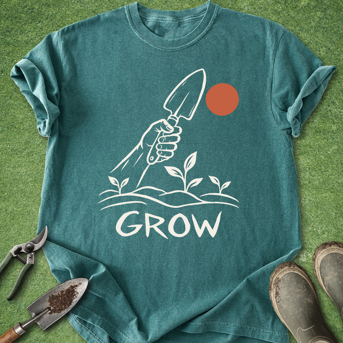 A teal short-sleeve t-shirt laid flat on a green grass-like surface, featuring a white graphic of a hand holding a trowel above small plants and the word 'GROW,' with an orange sun motif. Gardening tools and boots are placed around the shirt as styling props.