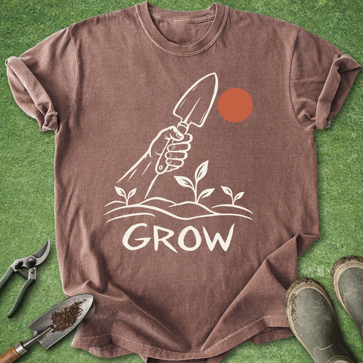 A brown short-sleeve t-shirt laid flat on green grass-like background, featuring a white hand holding a trowel above small plants and the word 'GROW,' with an orange circle above the plants. Gardening tools and muddy boots are placed around the shirt as styled props.