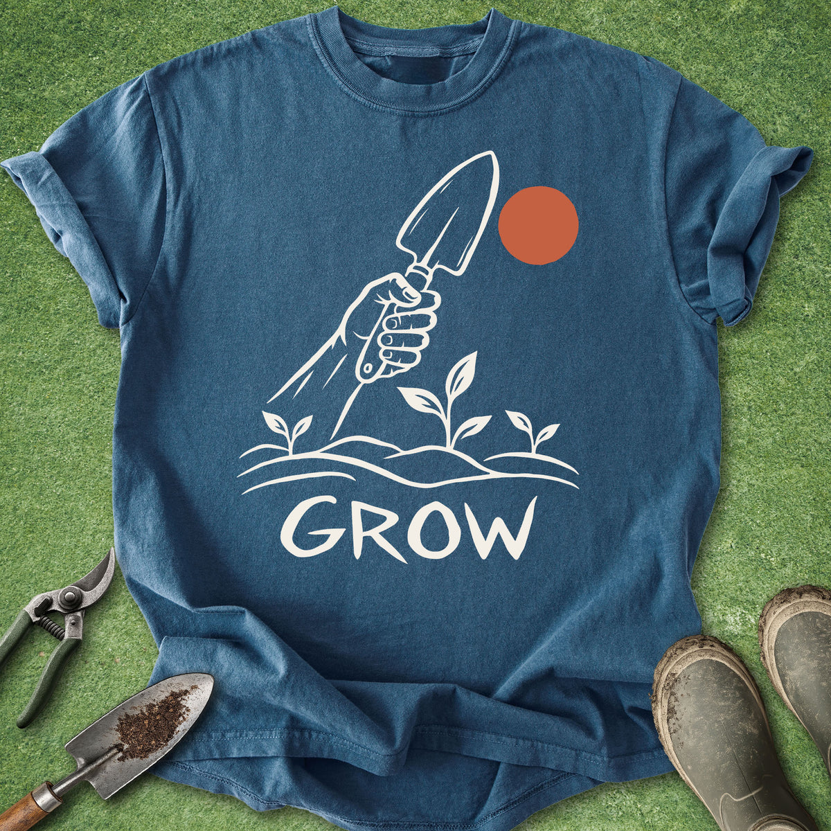 A blue short-sleeve graphic T-shirt laid flat on green grass with gardening props around it. The shirt has a white line drawing of a hand holding a trowel above small plants, with the word "GROW" in white below, and a small orange circle near the top right of the graphic.