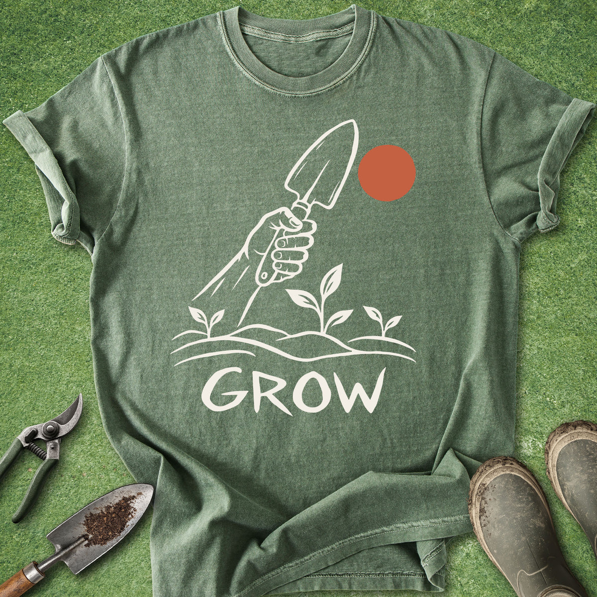A heathered sage green t-shirt with a gardening graphic, laid flat on a green grass-like background with gardening tools and boots as props.