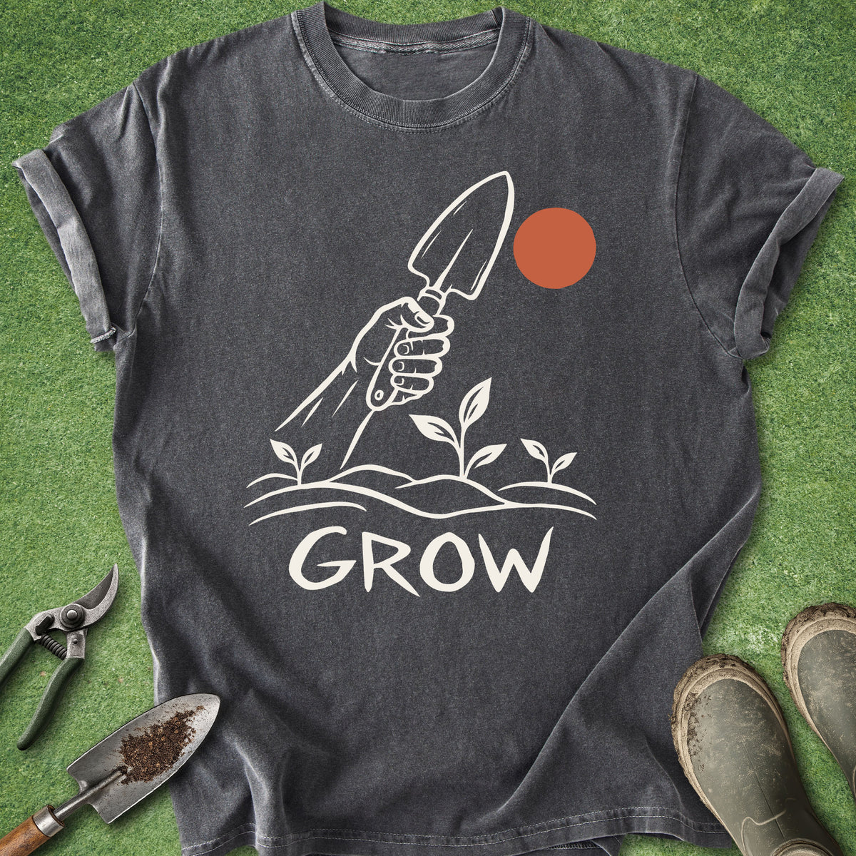 A dark gray graphic t-shirt laid flat on green grass with gardening props, including pruning shears, a trowel, and boots. The shirt features a white graphic of a hand holding a trowel above sprouting plants, with the word 'GROW' underneath and an orange sun circle.