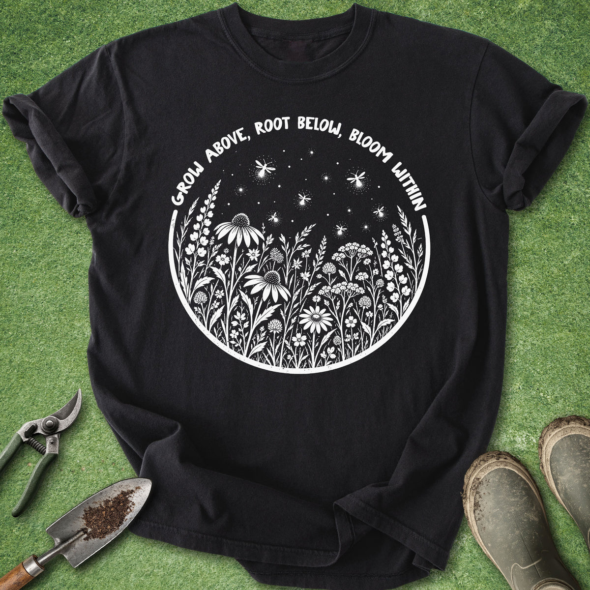 A black short-sleeve graphic T-shirt laid flat on green grass-like flooring, with gardening tools and boots around it. The shirt has a white circular floral design and the text 'GROW ABOVE, ROOT BELOW, BLOOM WITHIN'.