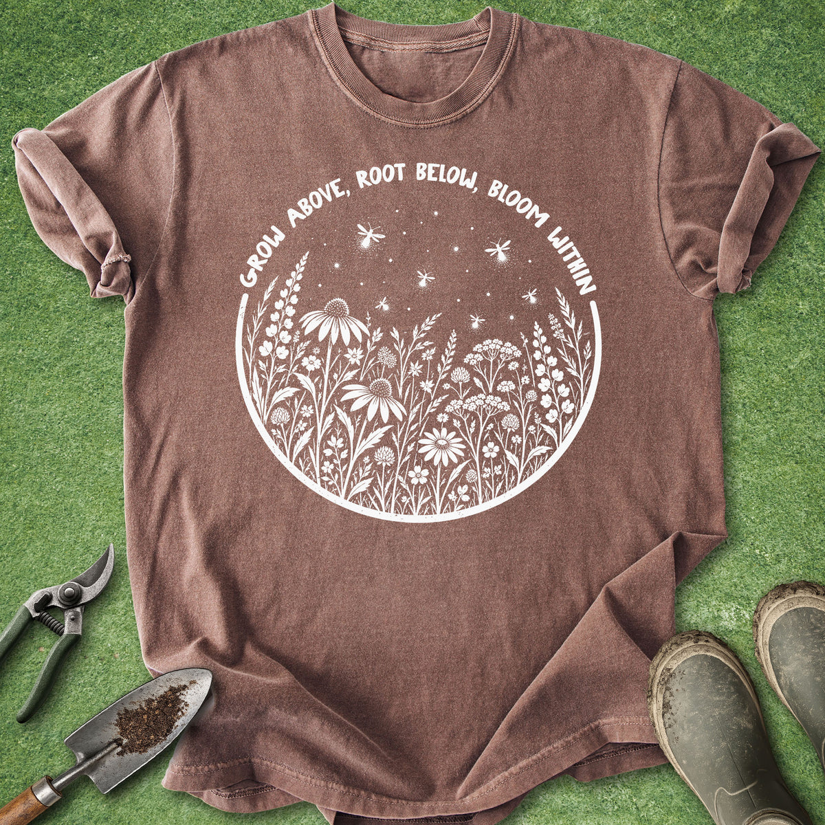 A brown short-sleeve graphic T-shirt laid flat on green grass-like background, with gardening tools and boots around it. The shirt has a white circular floral design and the text 'GROW ABOVE, ROOT BELOW, BLOOM WITHIN'.