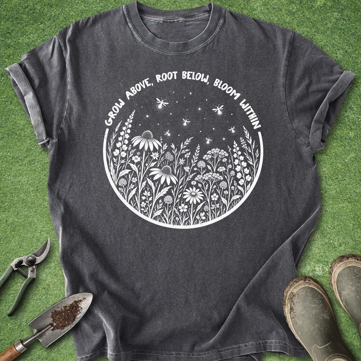 A dark gray t-shirt with a white circular graphic of wildflowers, stars, and the text 'GROW ABOVE, ROOT BELOW, BLOOM WITHIN', laid flat on green grass with gardening tools and boots nearby.