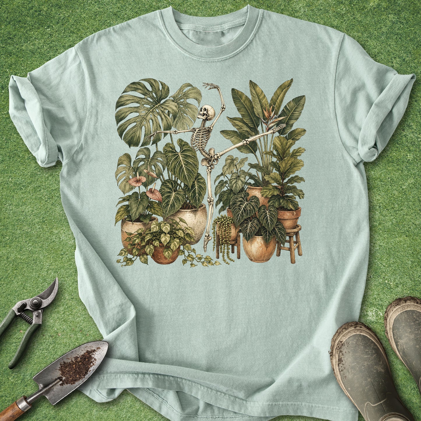A light mint green short-sleeve t-shirt laid flat on a green grass-like surface, featuring a vintage-style botanical illustration of a skeleton among potted houseplants. Gardening props including pruning shears, a trowel, and boots are placed around the shirt.