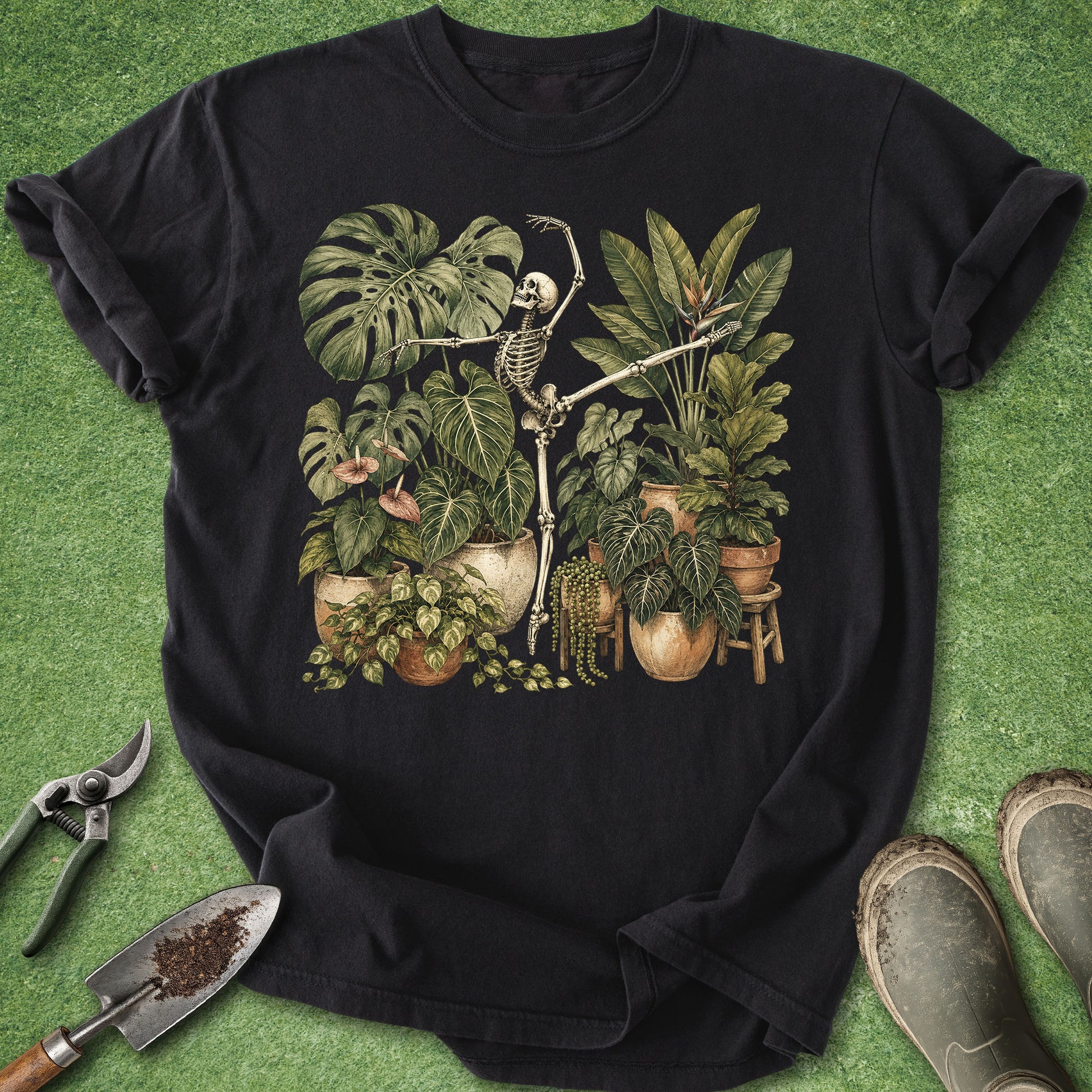 A black short-sleeve graphic T-shirt laid flat on a green textured surface, featuring a vintage-style illustration of a skeleton among potted houseplants. Gardening tools and boots are placed around the shirt as styling props.