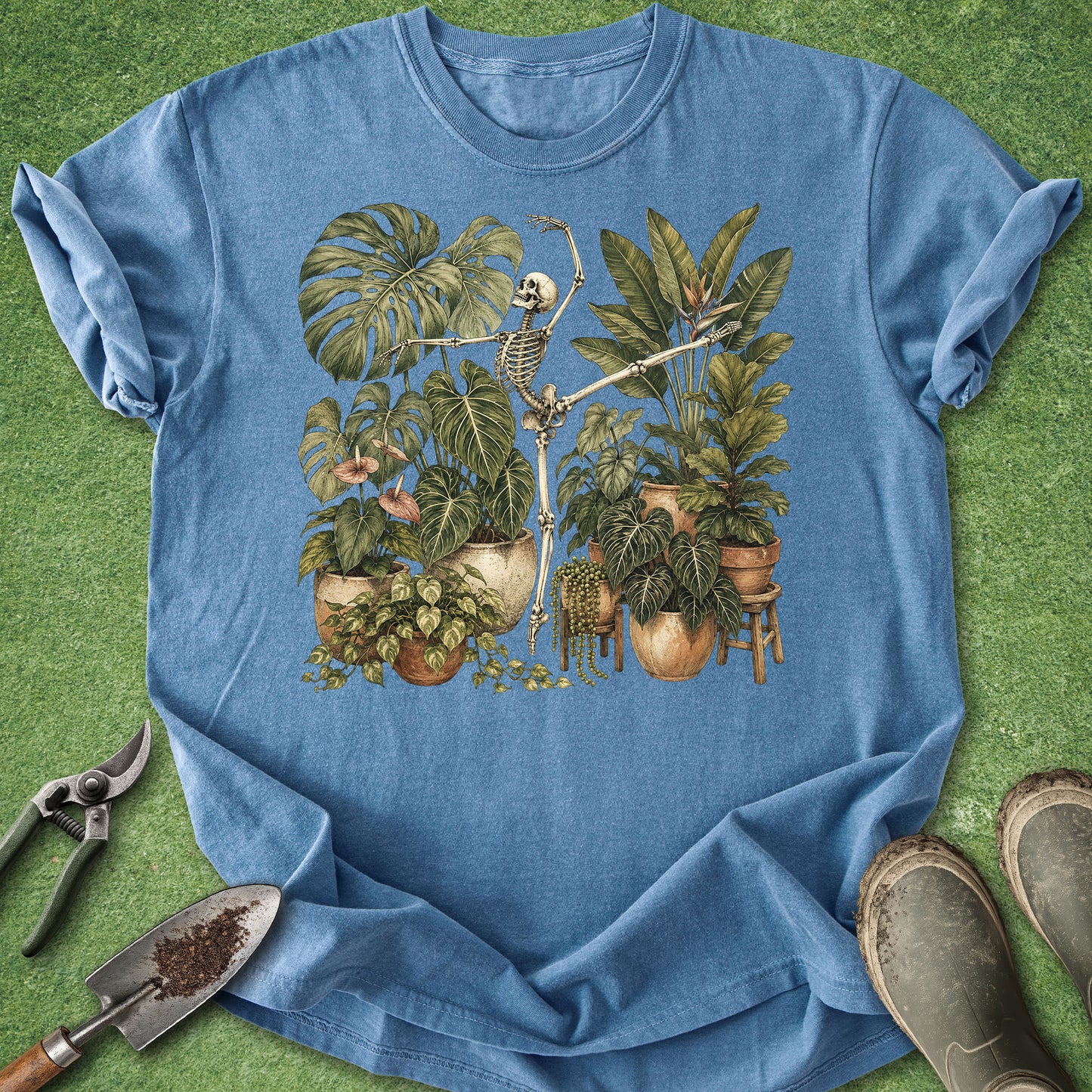 A blue short-sleeve T-shirt laid flat on a green grass-like surface, featuring a vintage-style illustration of a skeleton among potted tropical plants. Gardening tools and muddy boots are placed around the shirt as styling props.