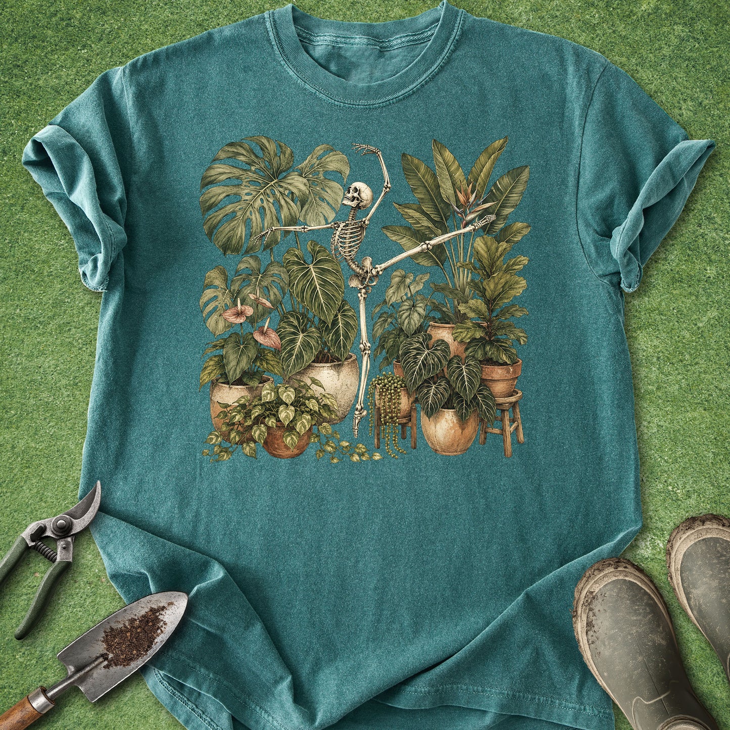 A teal short-sleeve t-shirt with a vintage-style graphic of a skeleton gardening among potted plants, laid flat on a green grass-like background with gardening tools and boots nearby.