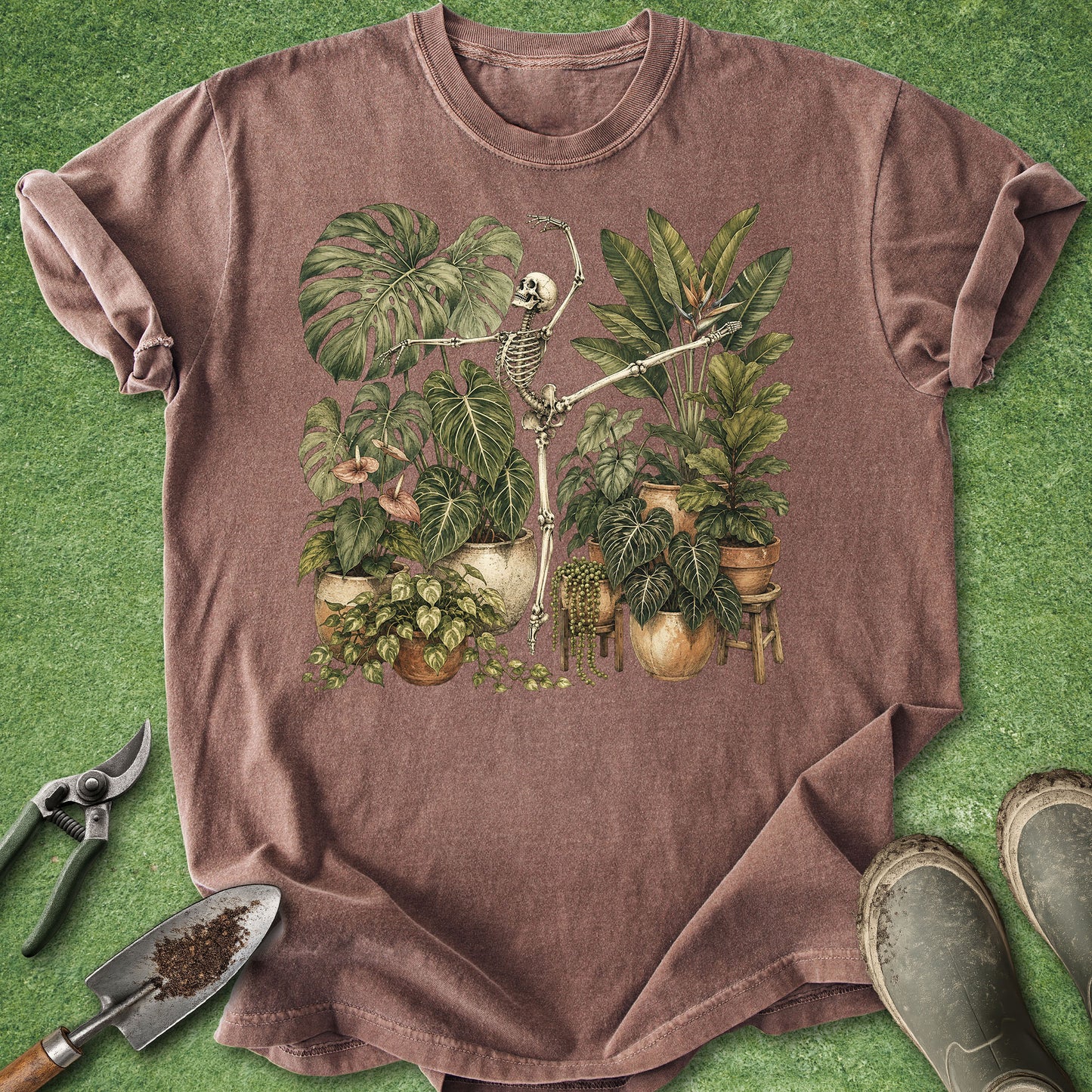A heathered brown short-sleeve t-shirt laid flat on green artificial turf, featuring a central graphic of a skeleton clock surrounded by potted tropical plants. Gardening props including pruning shears, a small hand fork with soil, and boots are arranged around the shirt.