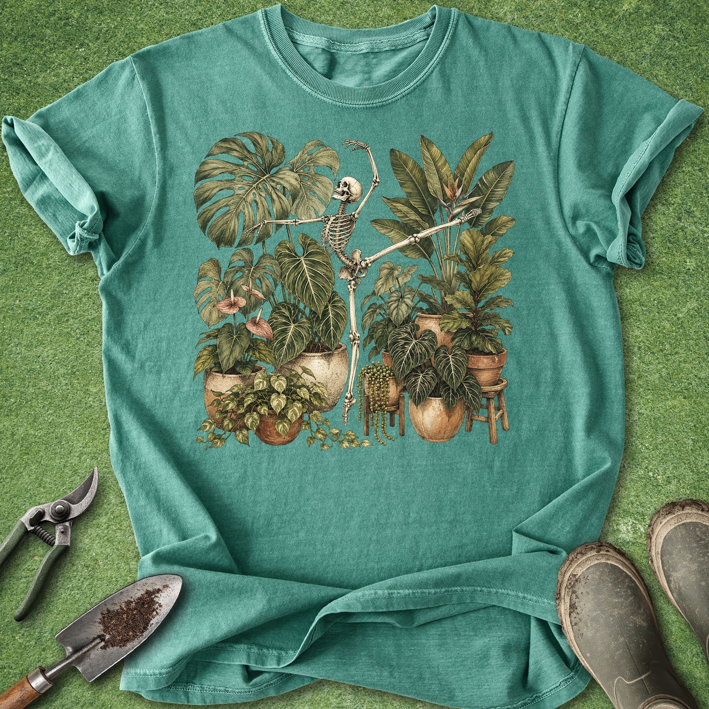 A teal short-sleeve crewneck t-shirt laid flat on a green grass-like surface, featuring a vintage-style graphic of a skeleton among potted tropical plants. Gardening props including pruning shears, a trowel, and boots are arranged around the shirt.