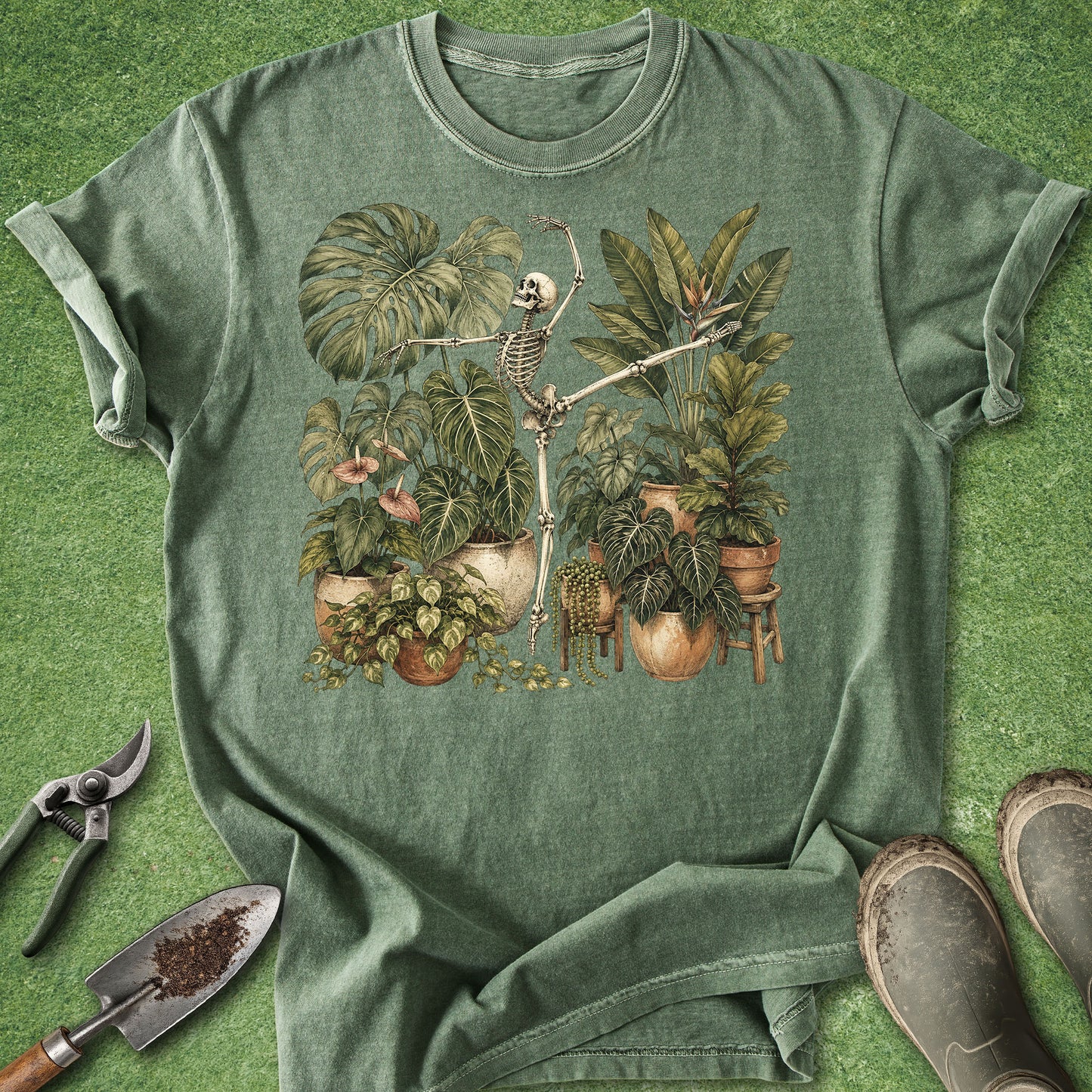 A sage green crewneck t-shirt featuring a vintage-style illustration of a skeleton gardening among potted plants, styled flat-lay on a green grass background with gardening tools and boots.