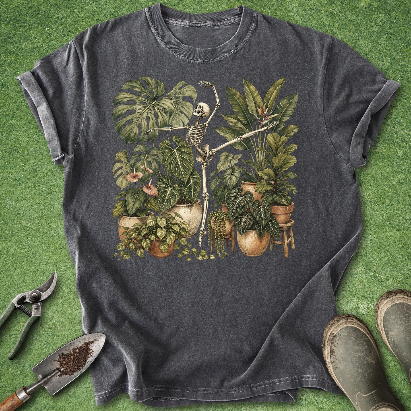 A dark charcoal gray t-shirt featuring a graphic of a skeleton clock surrounded by potted houseplants, styled in a flat-lay on artificial green turf with gardening tools and boots.