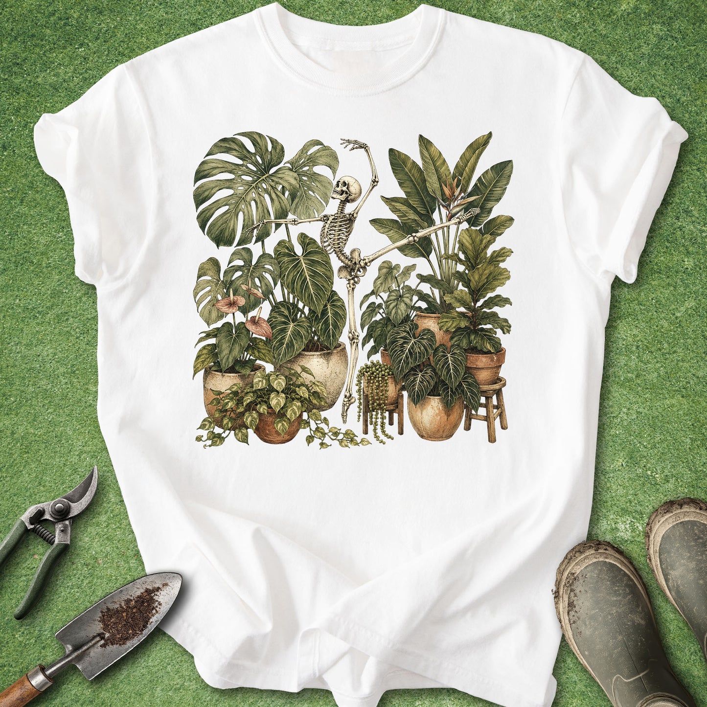 A white short-sleeve T-shirt laid flat on a green grass-like surface, featuring a large botanical illustration of potted houseplants and a skeleton on the front. Gardening props including pruning shears, a trowel, and boots are placed around the shirt.