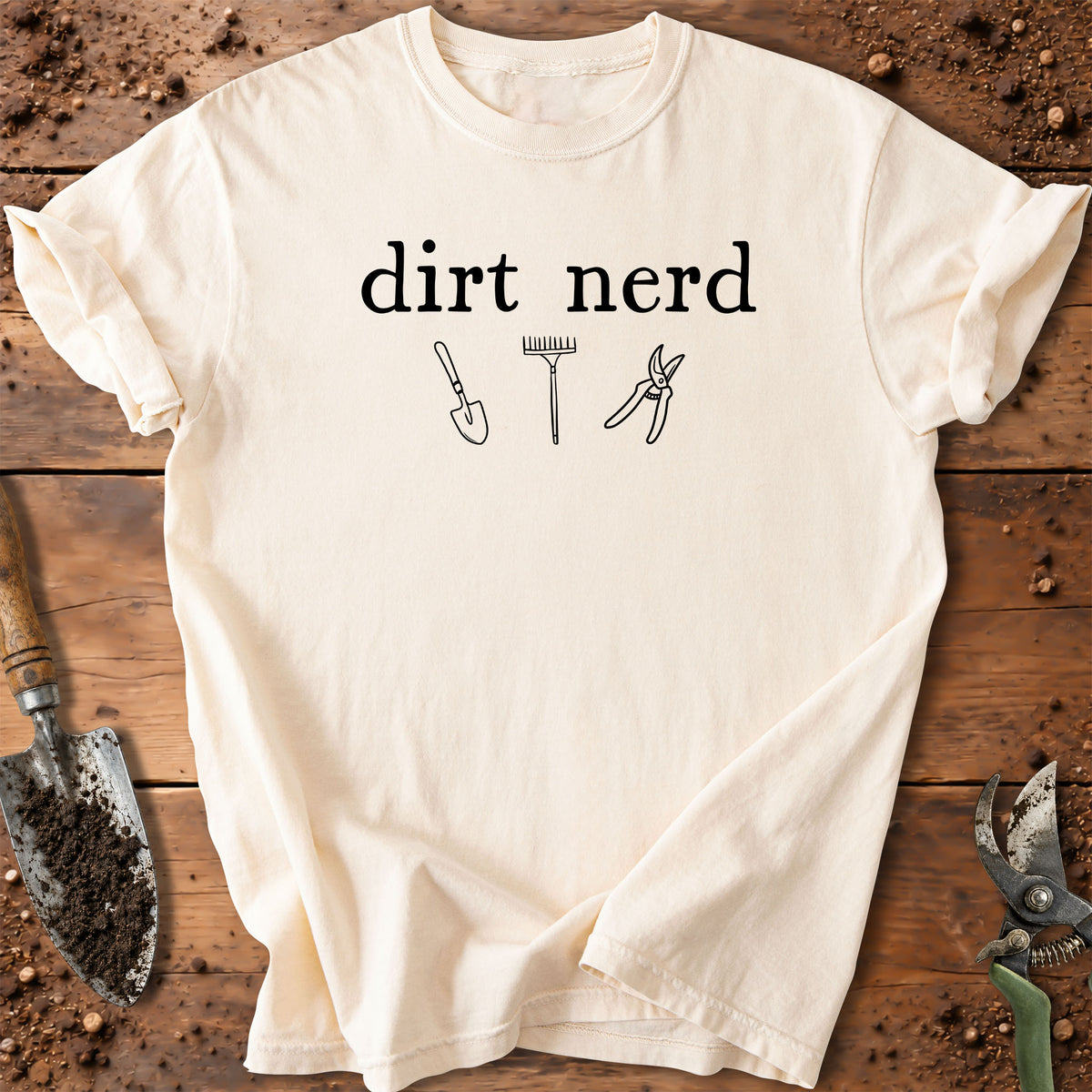 An off-white t-shirt with a black graphic reading 'dirt nerd' above simple line drawings of a shovel, rake, and pruning shears, laid flat on a rustic wooden surface with gardening tools around it.