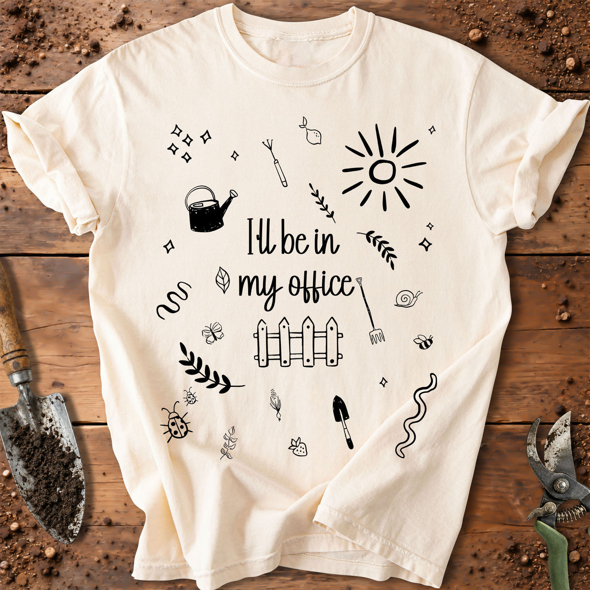 A cream-colored short-sleeve t-shirt with black hand-drawn garden-themed doodles and the text 'I'll be in my office' printed on the front, laid flat on a rustic wooden background with gardening tools.