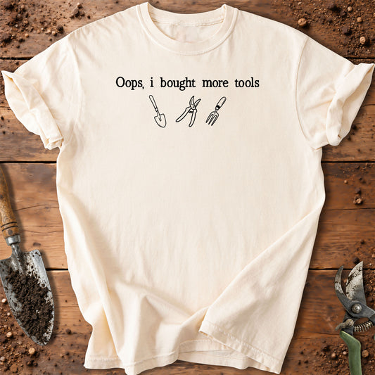 An off-white short-sleeve t-shirt with a black graphic print that reads 'Oops, i bought more tools' above simple line drawings of a trowel, pruning shears, and fork. The shirt is laid flat on a rustic wooden surface surrounded by gardening tools and soil.