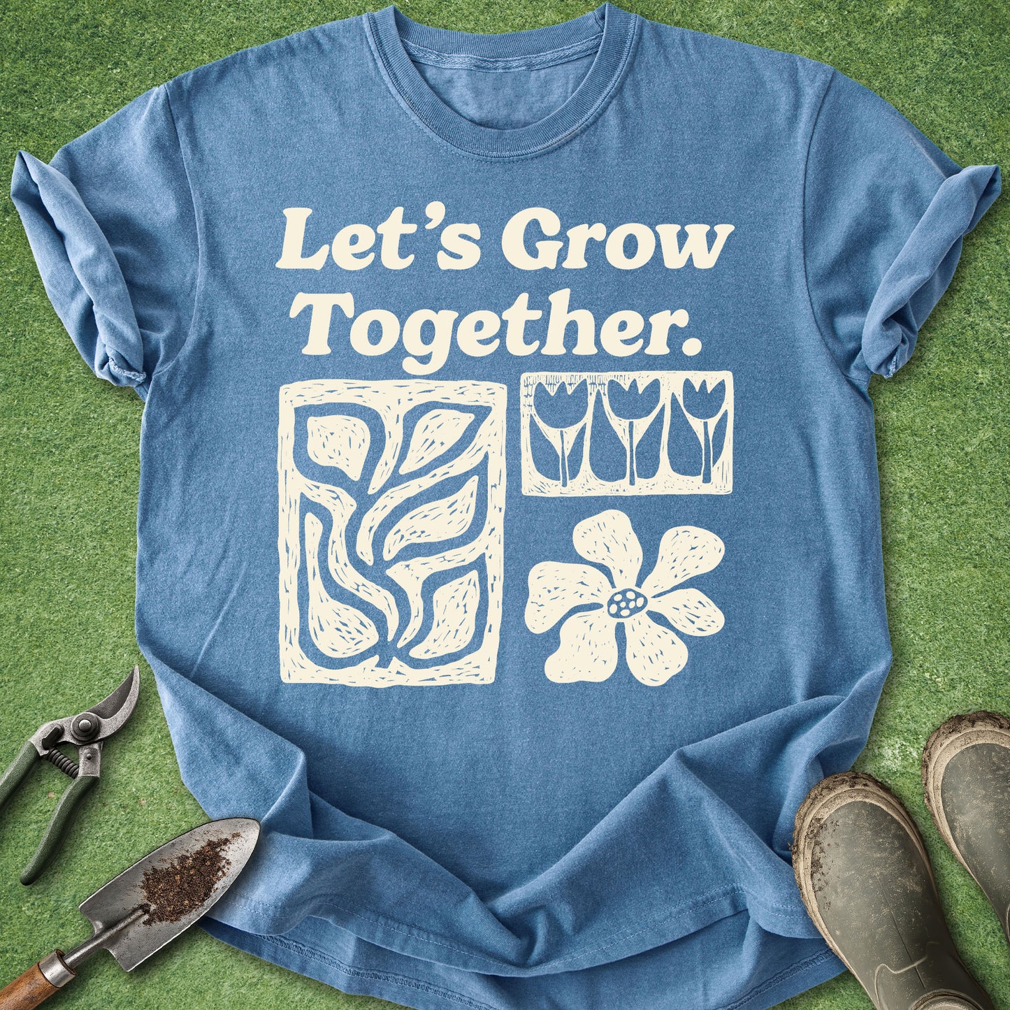 A blue short-sleeve graphic T-shirt laid flat on green grass-like ground, with gardening tools and boots around it. The shirt has white text reading 'Let's Grow Together.