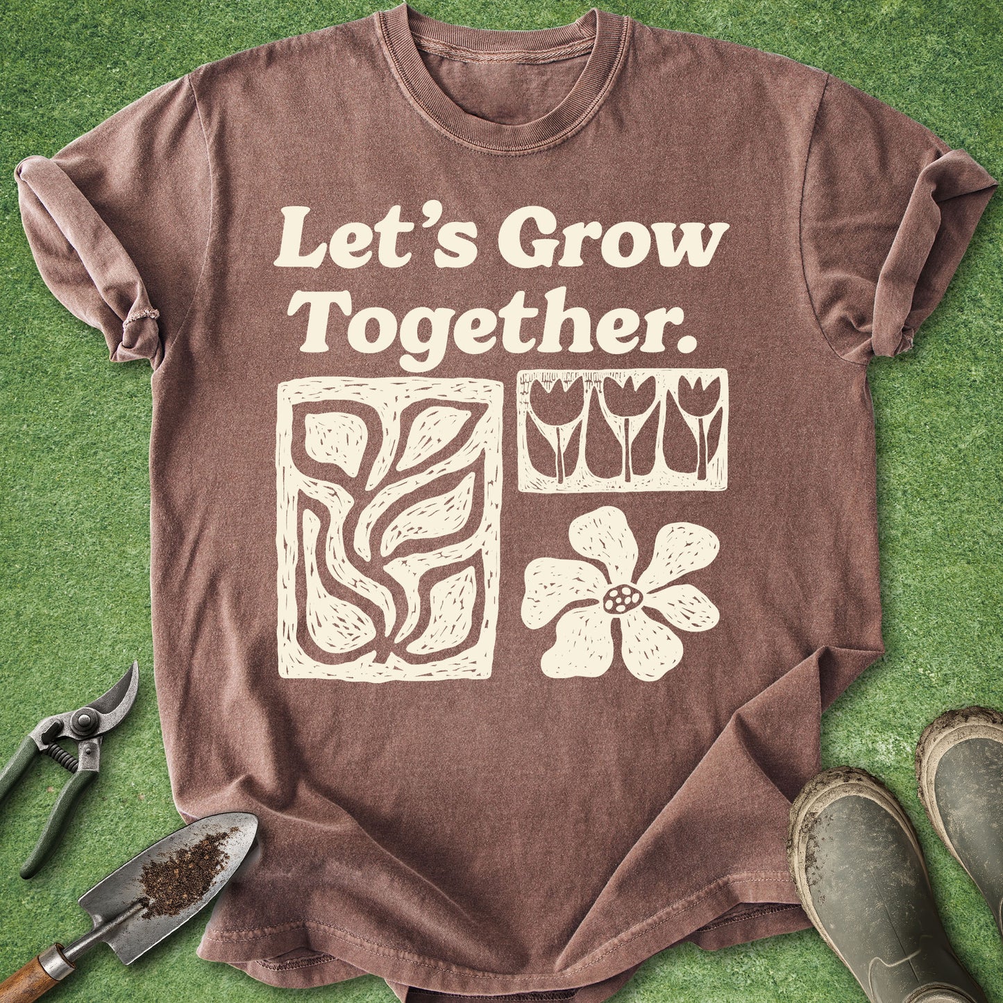 A brown short-sleeve graphic T-shirt laid flat on green grass-like background, with the phrase "Let's Grow Together." and simple plant illustrations printed in cream.