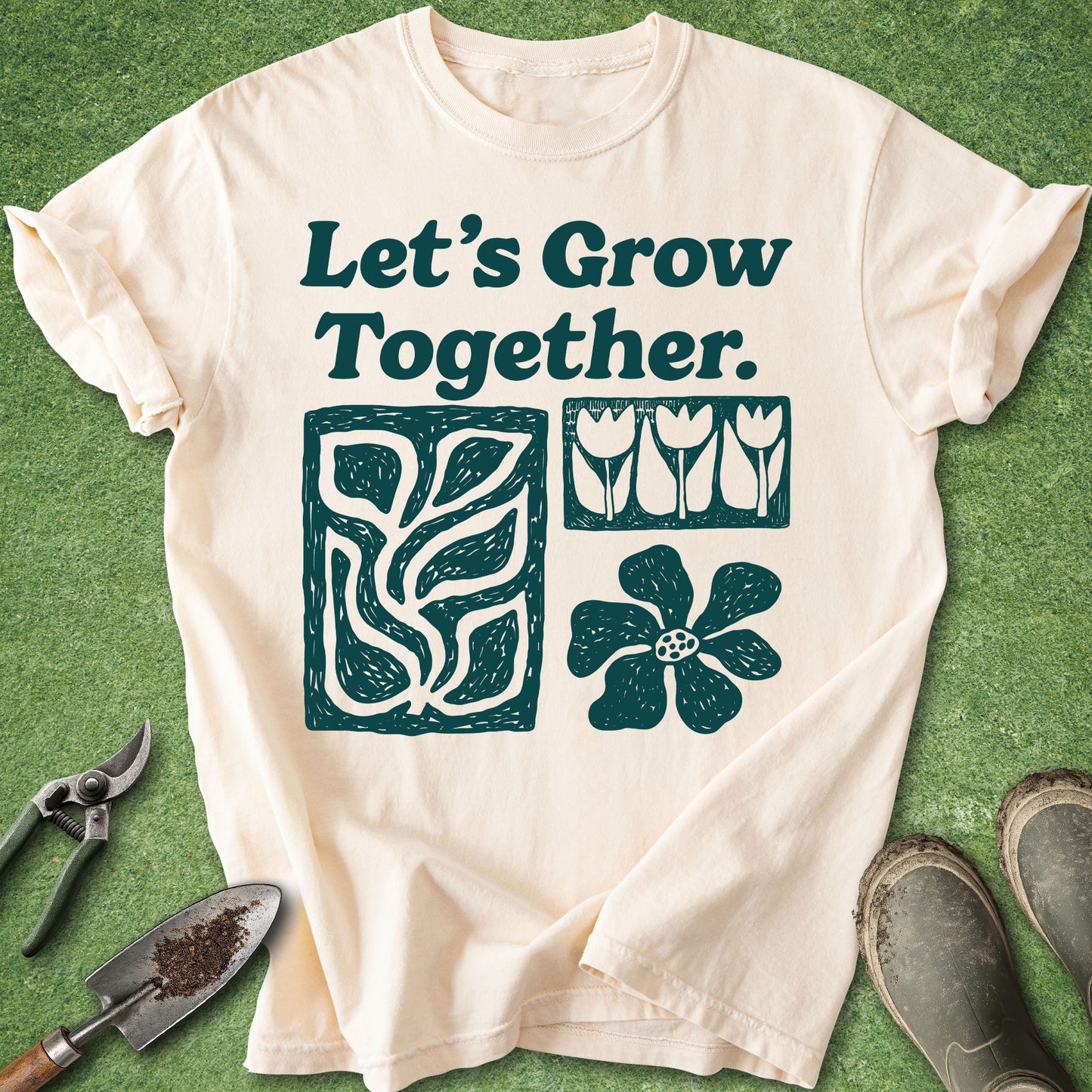 An off-white short-sleeve graphic T-shirt laid flat on green grass-like ground, with gardening tools and boots around it. The shirt reads “Let’s Grow Together.