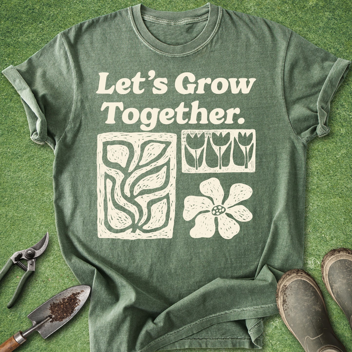 A green graphic t-shirt laid flat on a green grass-like surface, with gardening tools and boots around it. The shirt reads 'Let's Grow Together' above a leaf, tulips, and flower design.