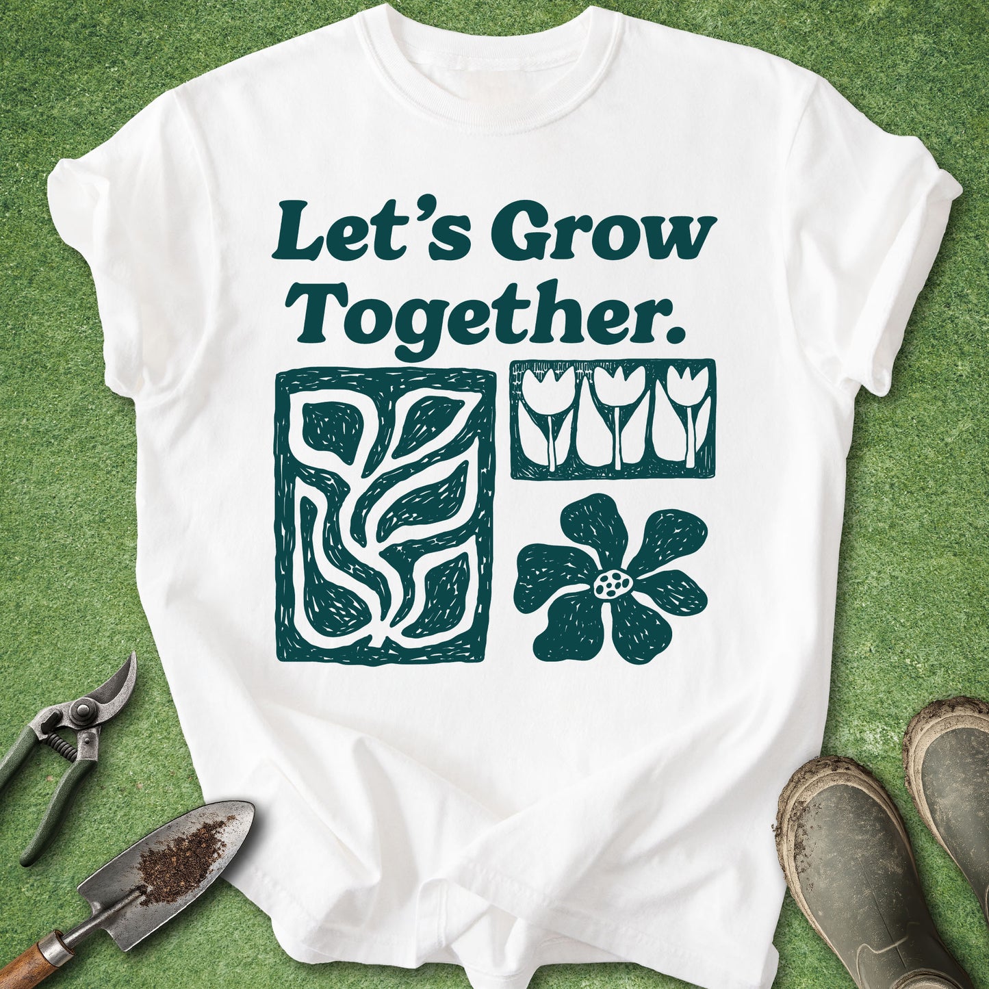 A white short-sleeve T-shirt laid flat on green grass with gardening props around it: pruning shears, a small hand trowel, and muddy boots. The shirt has dark teal text reading “Let’s Grow Together.