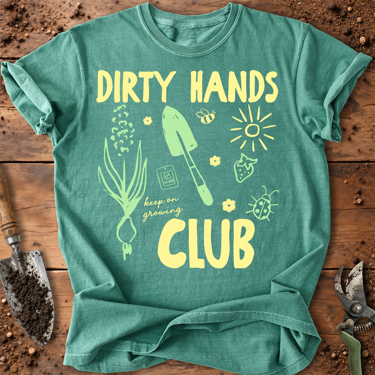 A green graphic t-shirt laid flat on a rustic wooden surface with gardening props like a trowel and pruning shears. The shirt features a yellow graphic that reads 'DIRTY HANDS CLUB' with illustrations of a plant, a trowel, a bee, a sun, and a ladybug.