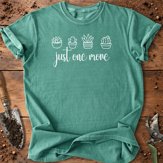 A light green short-sleeve crewneck t-shirt with a black graphic print of four potted cacti and the phrase 'just one more' in script, laid flat on a rustic wooden surface with gardening tools.