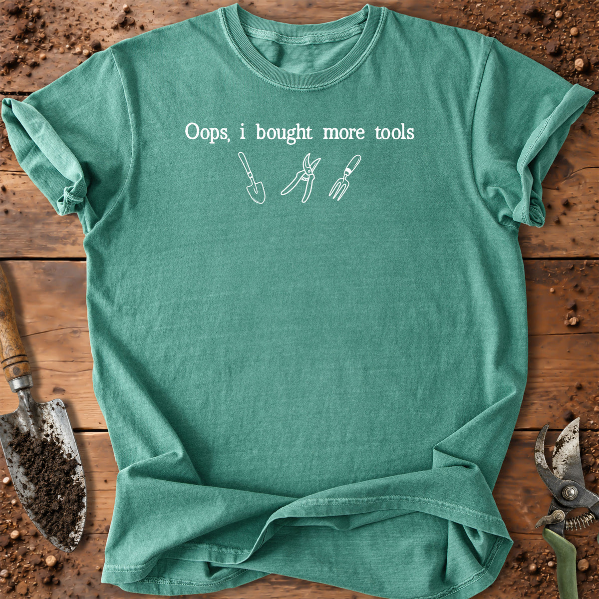 A green short-sleeve t-shirt with a white graphic print that reads 'Oops, i bought more tools' above simple drawings of gardening tools, laid flat on a rustic wooden surface with soil and tools around it.