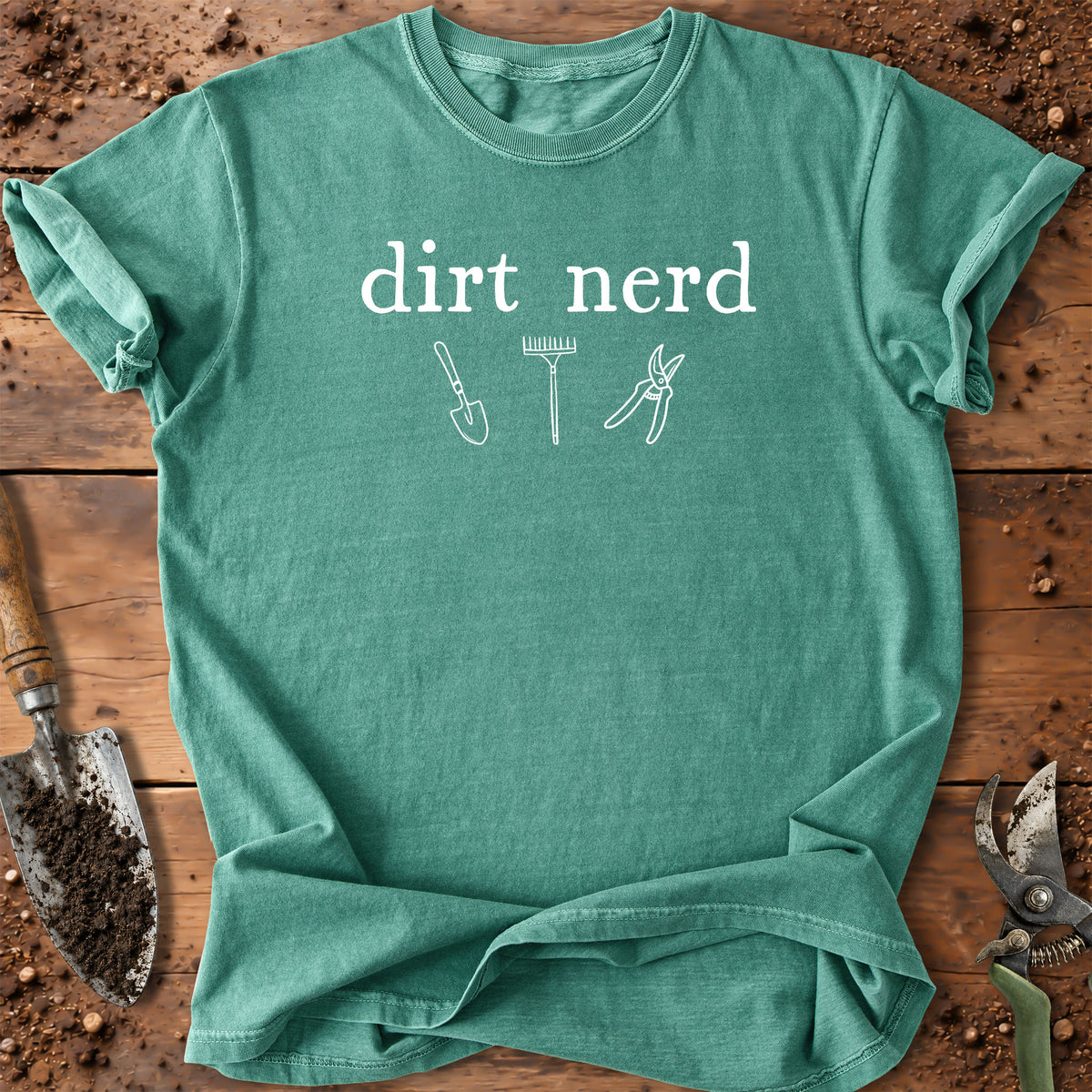 A heathered teal green t-shirt with a white graphic that reads 'dirt nerd' above simple line drawings of gardening tools (trowel, rake, pruner). The shirt is laid flat on a rustic wooden background surrounded by gardening props like a trowel and shears.