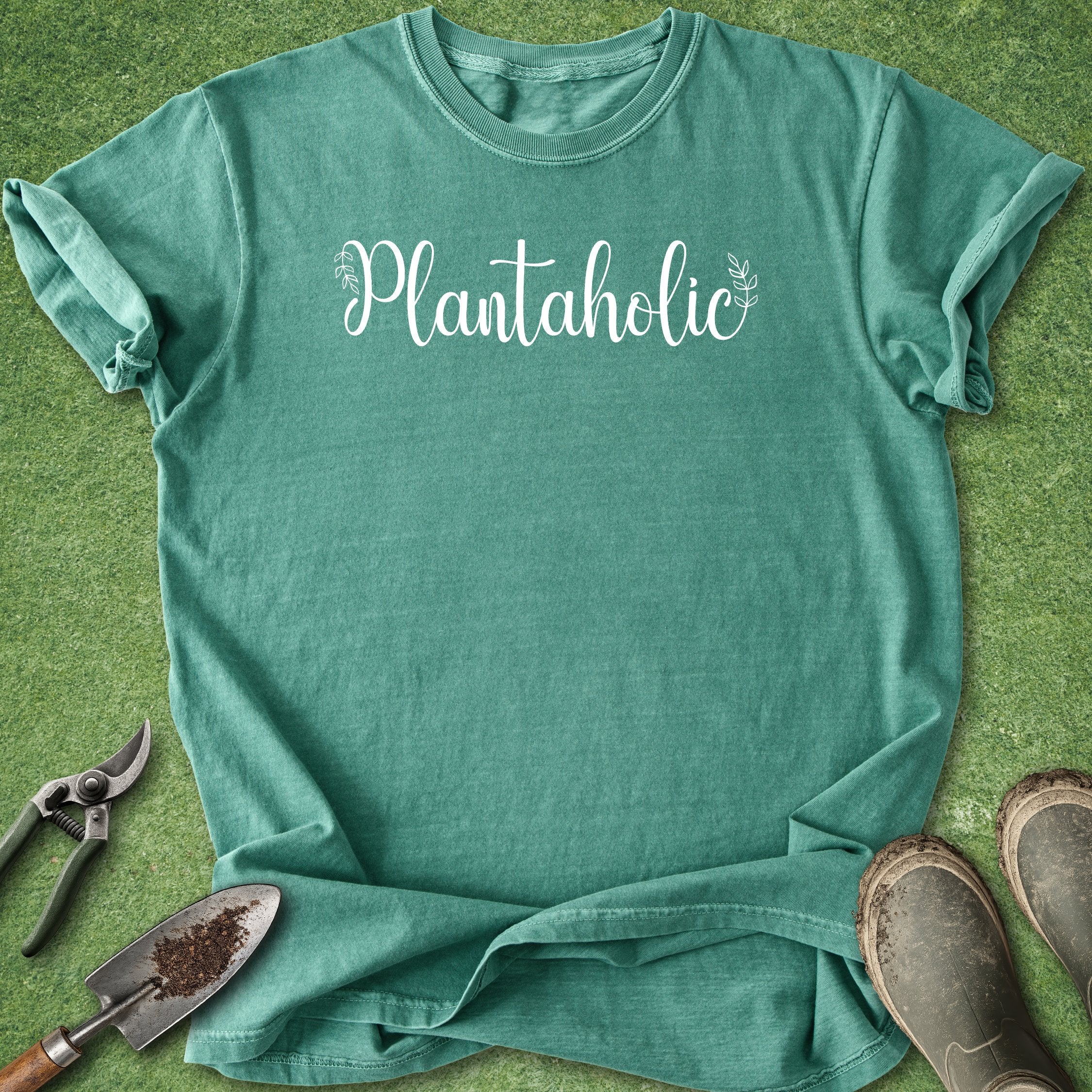 Green t-shirt with 'Plantaholic' text on a grass background with gardening tools.