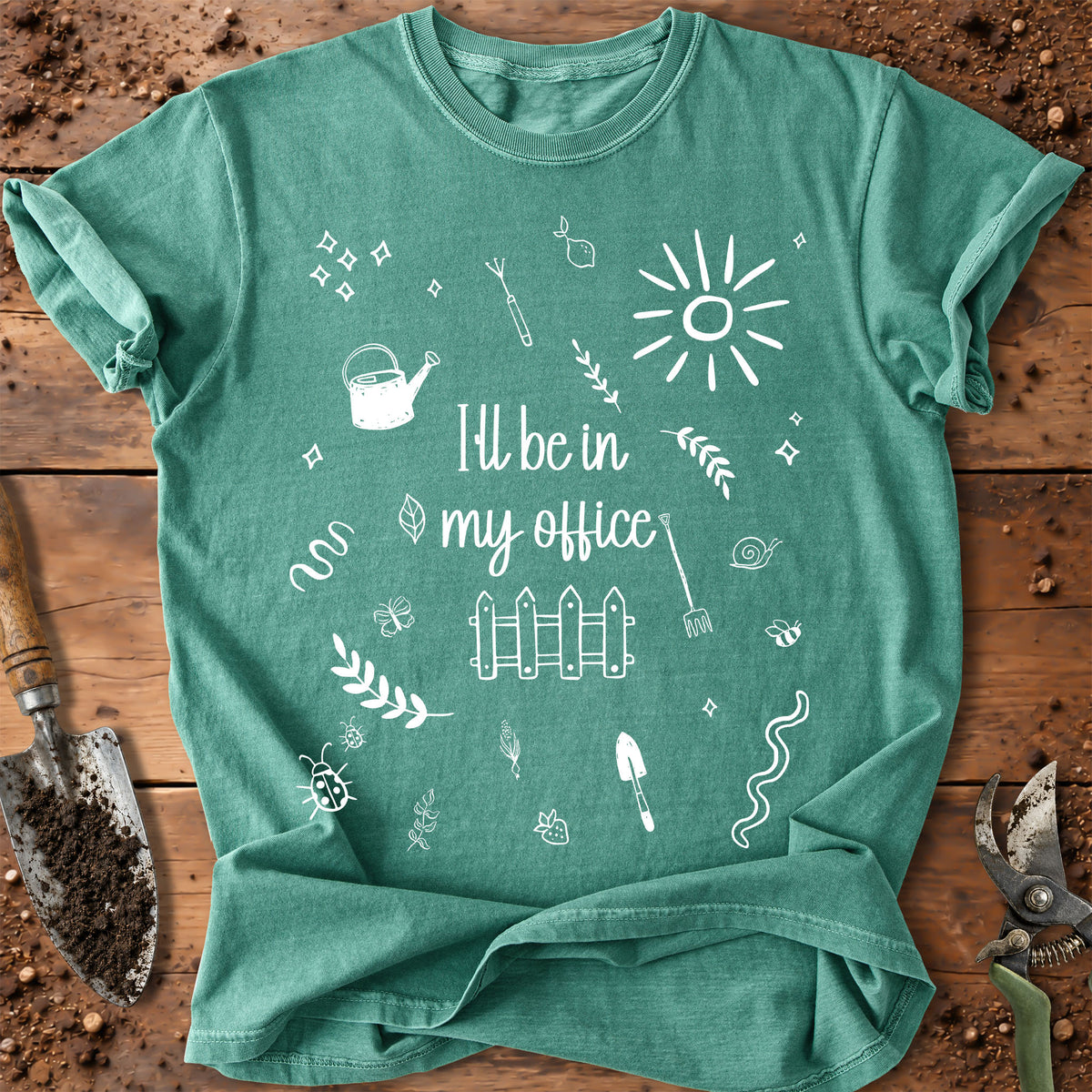 A green graphic t-shirt laid flat on a rustic wooden surface with gardening tools around it. The shirt features a white hand-drawn style print of a sun, fence, watering can, and the phrase 'I'll be in my office'.