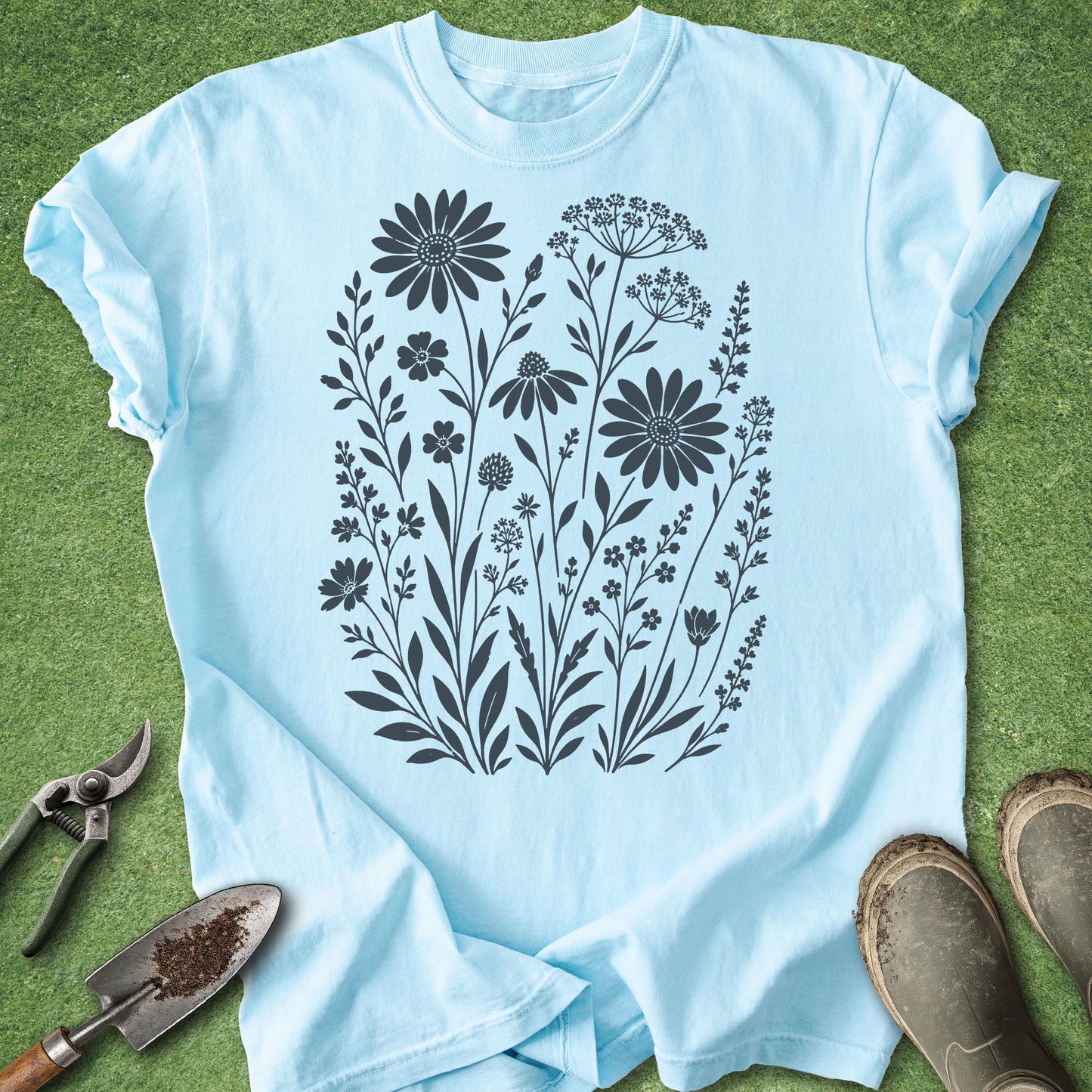 A light blue short-sleeve T-shirt laid flat on green grass, featuring a large black floral print on the front. Gardening tools and muddy boots are placed around the shirt as styling props.