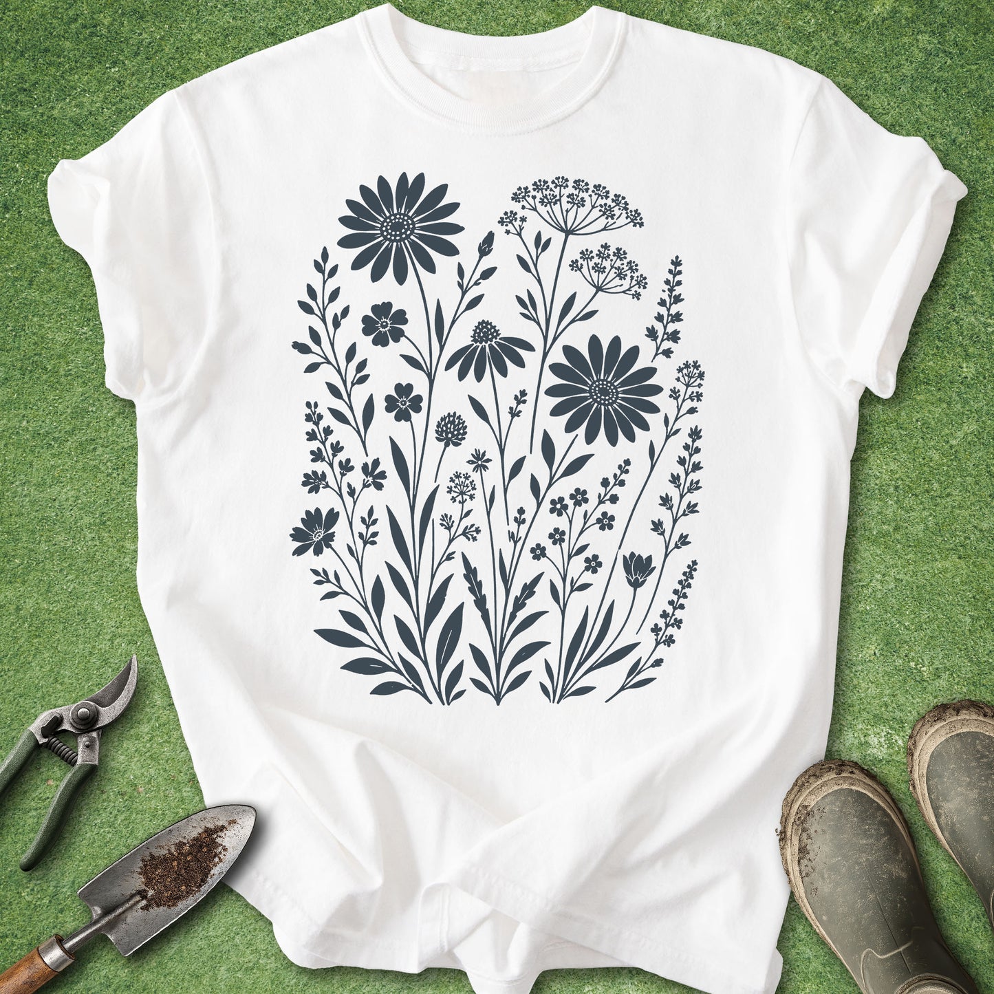 A white short-sleeve T-shirt laid flat on green artificial grass, featuring a dark navy floral print of wildflowers centered on the chest. Gardening props including pruning shears, a trowel, and boots are placed around the shirt.