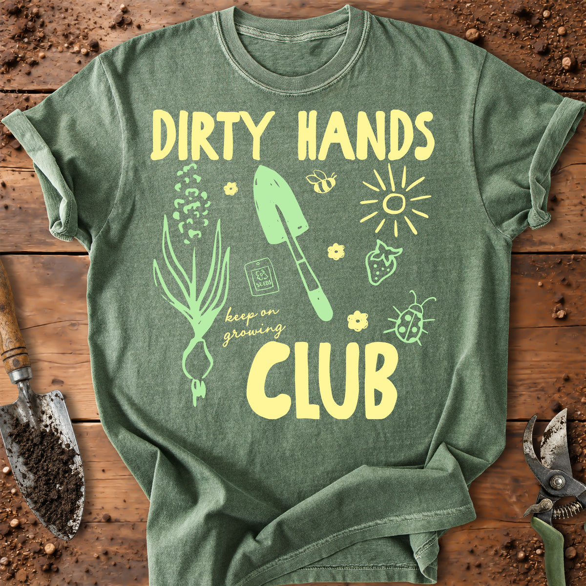 A green graphic t-shirt laid flat on a rustic wooden surface with gardening tools. The shirt features a yellow and light green graphic that reads 'DIRTY HANDS CLUB' with illustrations of a garden sprout, trowel, sun, strawberry, bee, and ladybug.