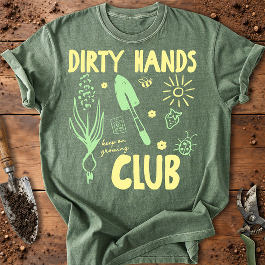 A green graphic t-shirt laid flat on a rustic wooden surface with gardening tools. The shirt features a yellow and light green graphic that reads 'DIRTY HANDS CLUB' with illustrations of a garden sprout, trowel, sun, strawberry, bee, and ladybug.