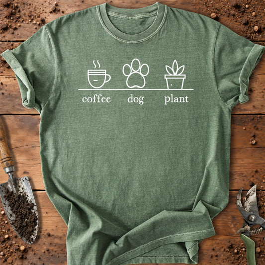 A heathered sage green t-shirt with a white graphic print of a coffee cup, a dog paw print, and a potted plant labeled 'coffee', 'dog', and 'plant'. The shirt is laid flat on a rustic wooden background with gardening props like a trowel and pruning shears.