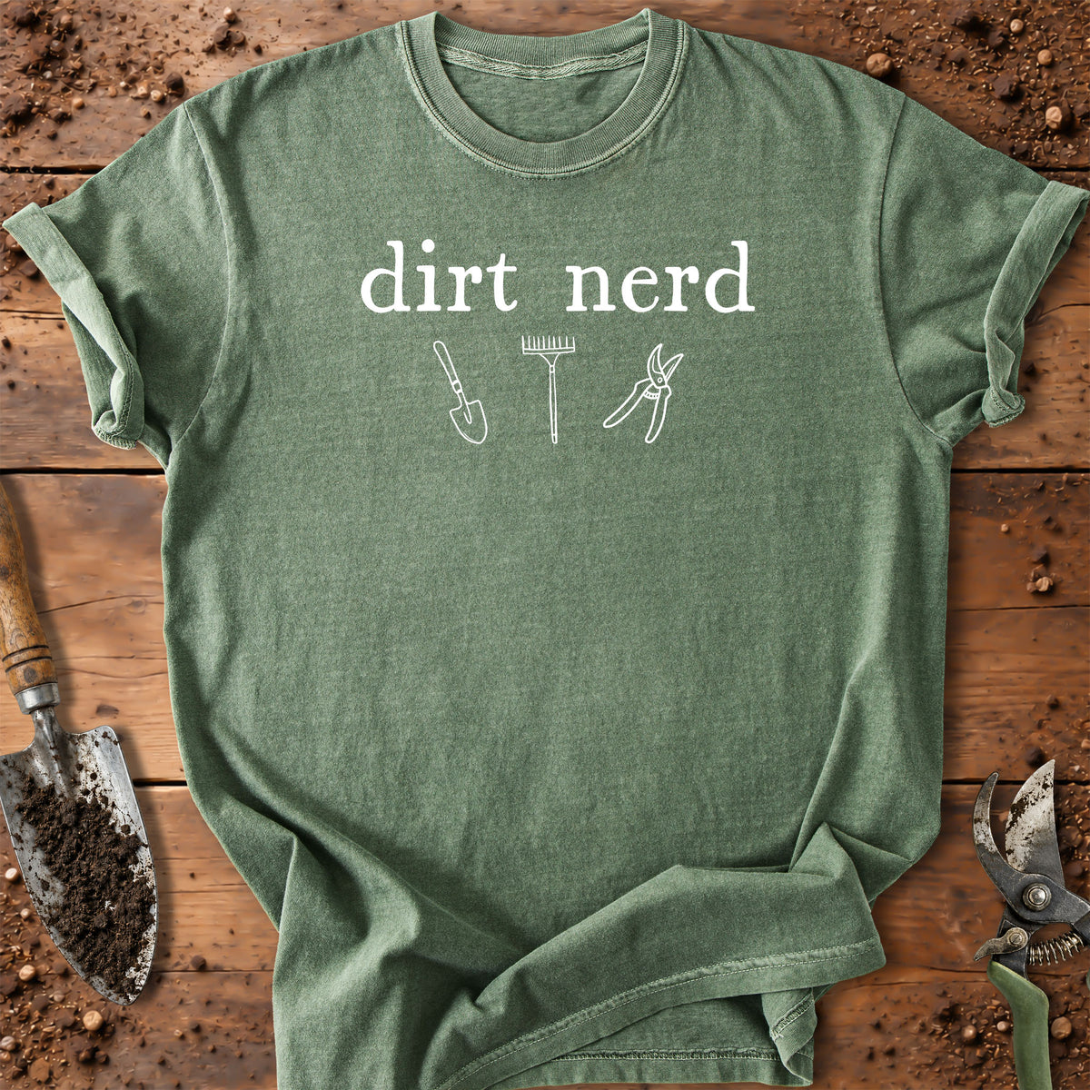 A green short-sleeve t-shirt with a white graphic that reads 'dirt nerd' above simple line drawings of gardening tools, laid flat on a rustic wooden surface with soil and gardening tools around it.