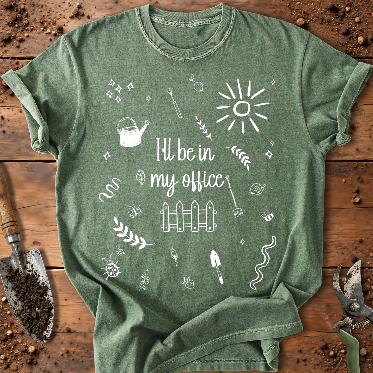 A green graphic t-shirt laid flat on a rustic wooden background with gardening tools. The shirt features a white hand-drawn style print that reads 'I'll be in my office' surrounded by doodles of a sun, fence, watering can, and gardening tools.