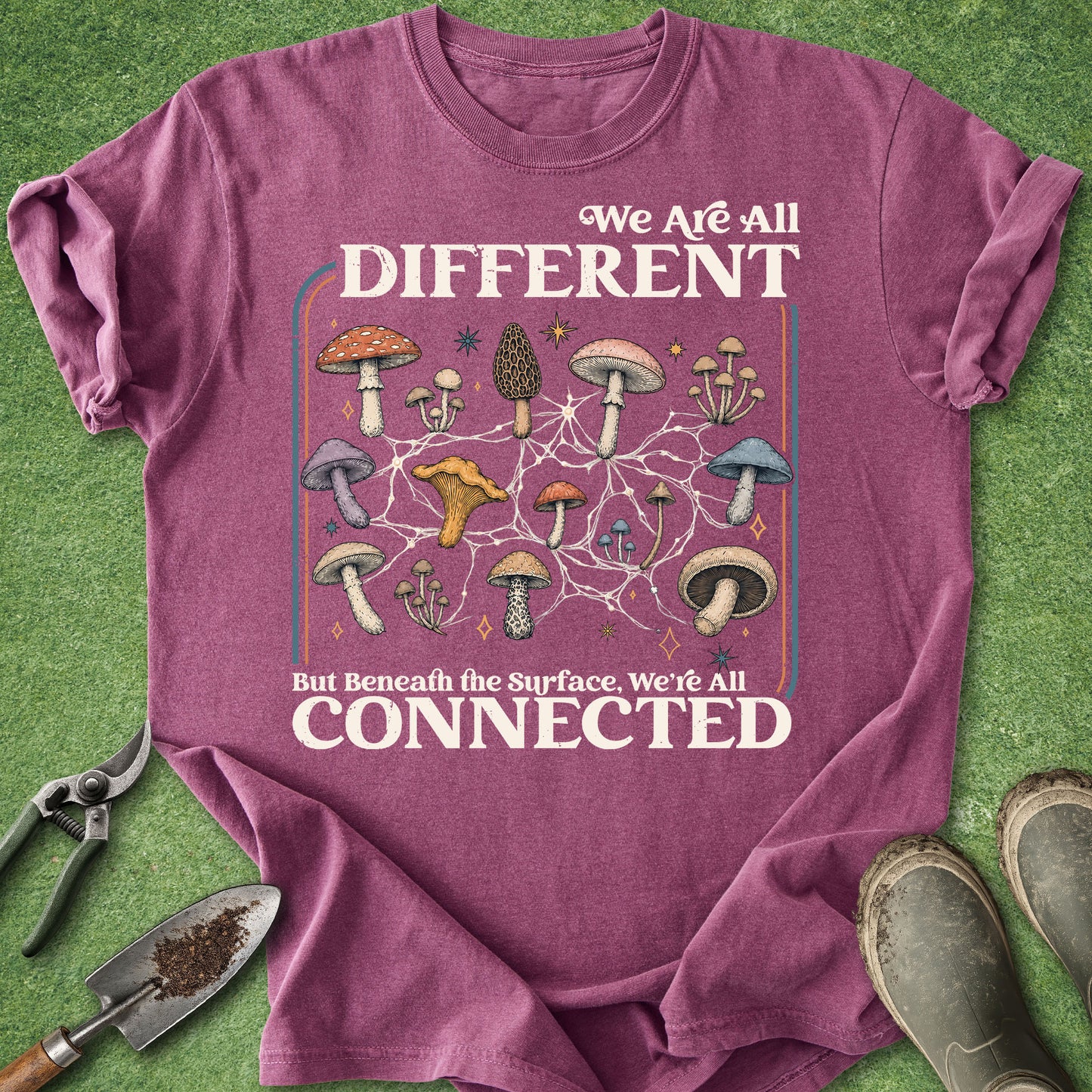 A heathered purple graphic t-shirt laid flat on a green grass-like surface, with gardening tools and boots around it. The shirt features a mushroom network illustration and the text 'We Are All DIFFERENT But Beneath the Surface, We're All CONNECTED'.