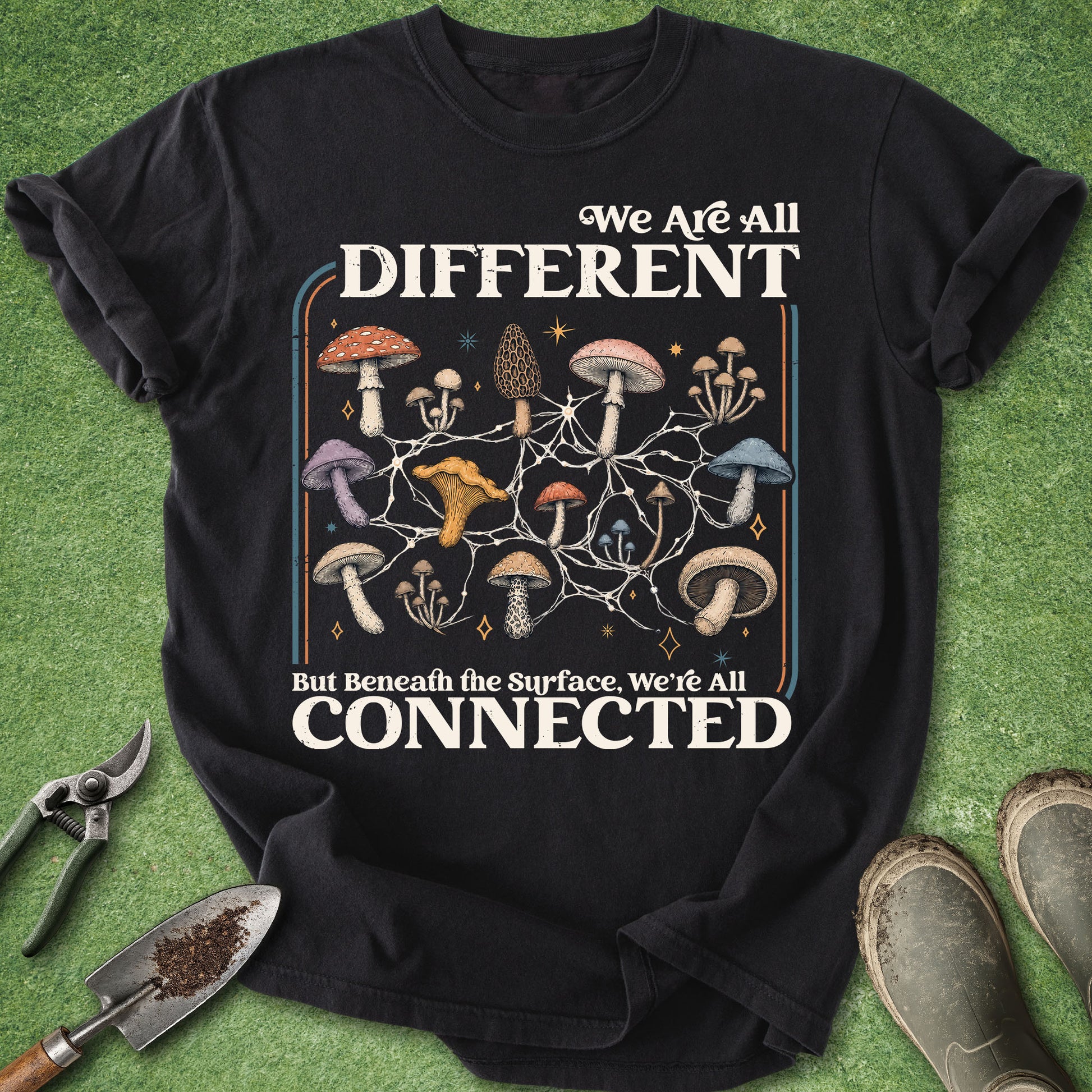 A black graphic t-shirt laid flat on green grass with gardening tools and boots around it. The shirt features a mushroom illustration and the text 'We Are All DIFFERENT But Beneath the Surface, We're All CONNECTED'.