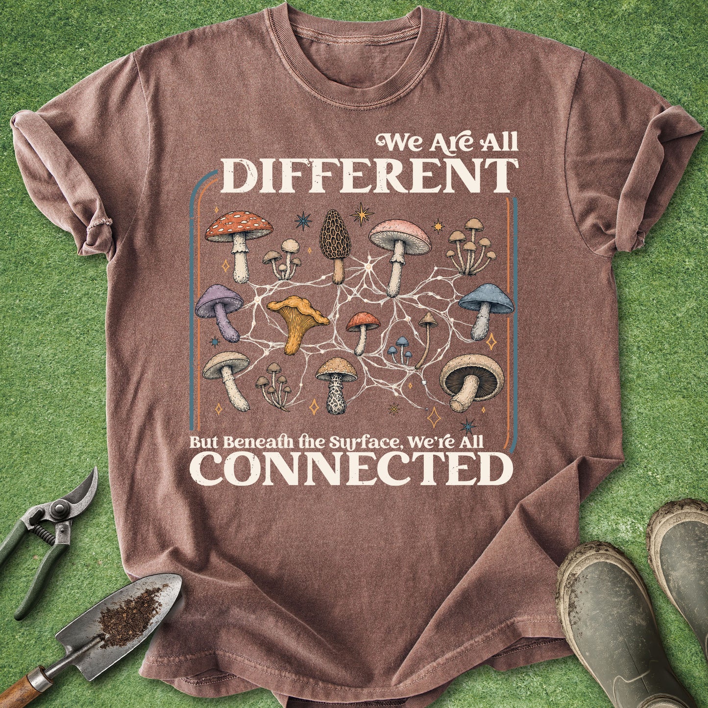 A brown graphic t-shirt laid flat on green grass with gardening tools and boots around it. The shirt features a mushroom network design with the text 'We Are All DIFFERENT But Beneath the Surface, We're All CONNECTED'.
