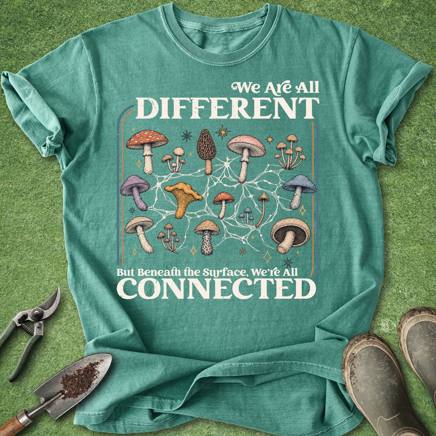 A teal graphic t-shirt laid flat on a green grass-like surface, with gardening tools and boots around it. The shirt has a large mushroom illustration and the text 'We Are All DIFFERENT But Beneath the Surface, We're All CONNECTED'.