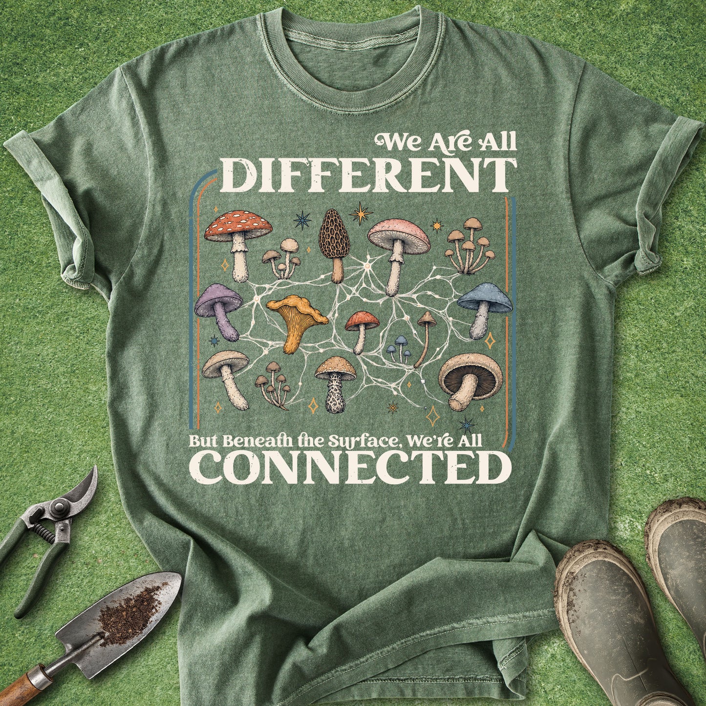 A sage green graphic t-shirt laid flat on a textured green grass background, styled with gardening tools (trowel, pruning shears) and muddy boots. The t-shirt features a detailed graphic of various illustrated mushrooms connected by a network diagram, with the text 'We Are All DIFFERENT But Beneath the Surface, We're All CONNECTED'.