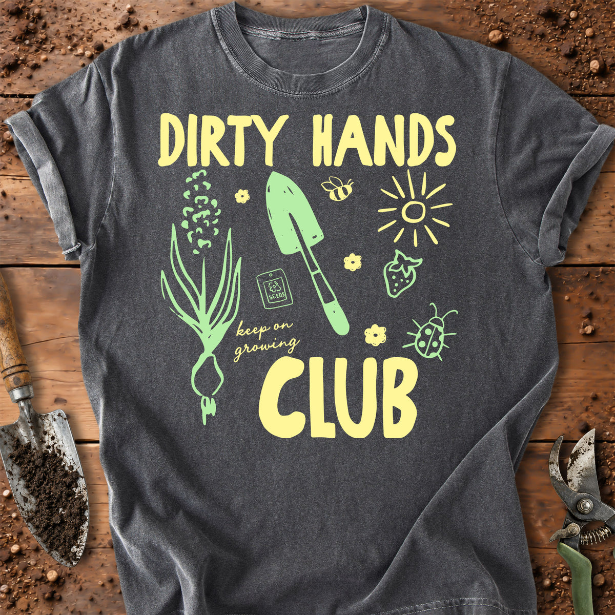 A dark gray graphic t-shirt laid flat on a rustic wooden surface with gardening props like a trowel, seed, and pruning shears. The shirt features a yellow and green graphic design with the text 'DIRTY HANDS CLUB' and illustrations of a plant, trowel, bee, sun, strawberry, and ladybug.