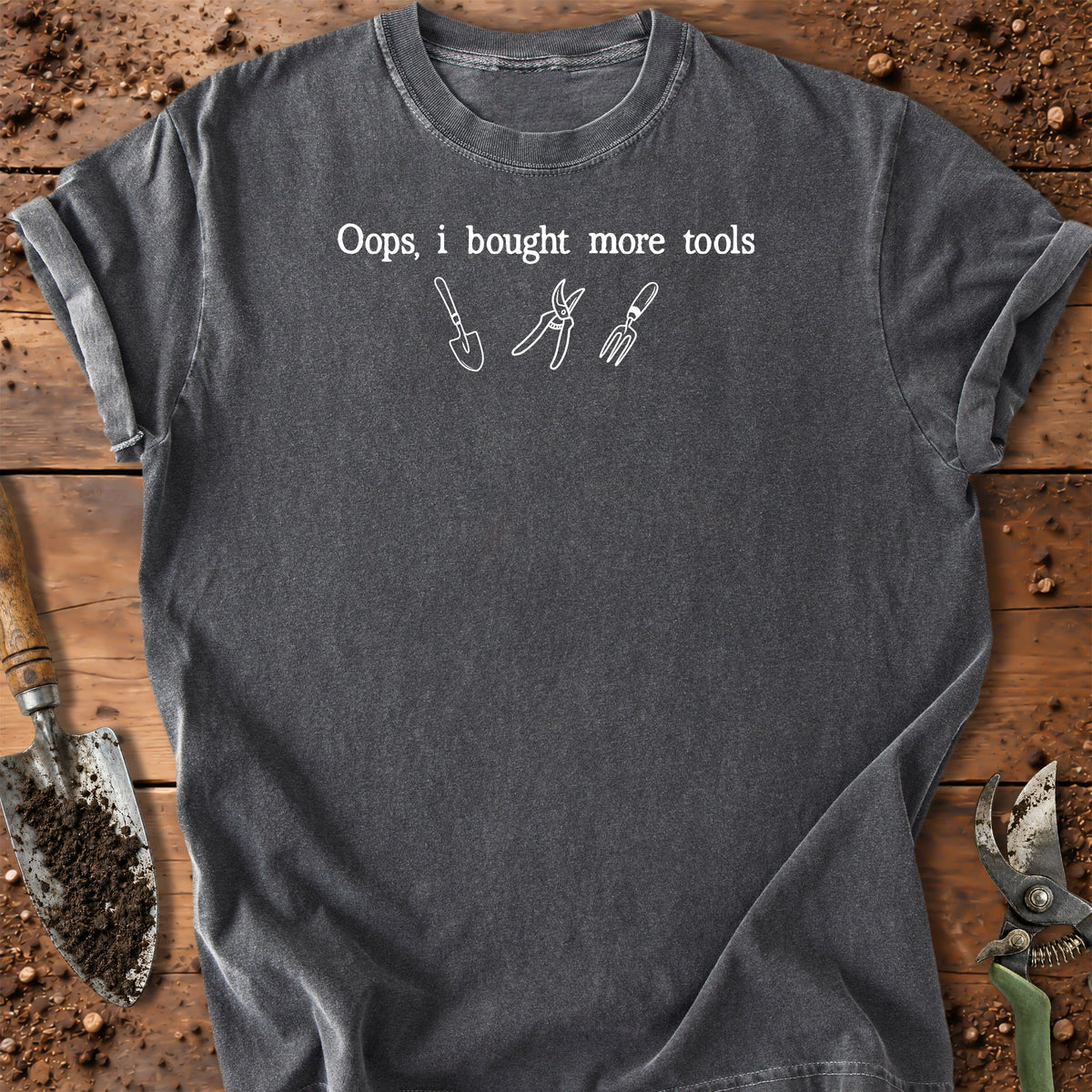 A dark gray t-shirt laid flat on a rustic wooden surface with gardening tools around it. The shirt features the white printed text 'Oops, i bought more tools' above simple white line drawings of a trowel, pruning shears, and a fork.