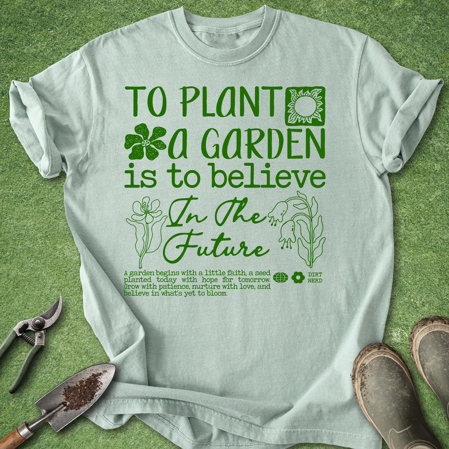 A light sage green graphic t-shirt laid flat on a green grass-like surface, with gardening tools and shoes around it. The shirt features a green printed quote that reads 'TO PLANT A GARDEN IS TO BELIEVE In The Future' with small plant illustrations.