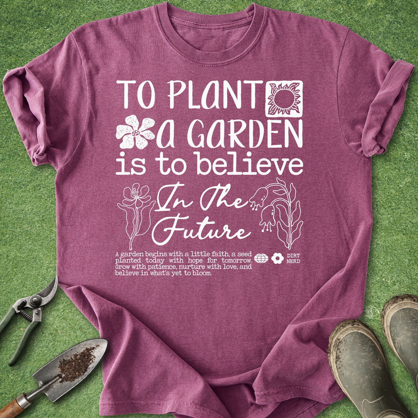 A mauve-purple short-sleeve graphic t-shirt laid flat on a green grass-like surface, with gardening tools and boots around it. The shirt has a white printed message reading 'TO PLANT A GARDEN IS TO BELIEVE In The Future' with floral and plant illustrations.