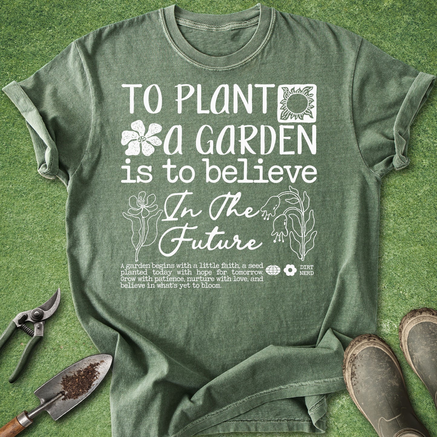An olive green graphic t-shirt laid flat on a green grass-like surface, surrounded by gardening props including pruning shears, a trowel, and boots. The shirt features a white printed design with the text 'TO PLANT A GARDEN IS TO BELIEVE In The Future' along with floral and plant illustrations.